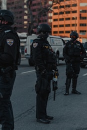 A group of police officers in tactical gear is standing on a city street. They are wearing black uniforms, helmets, and protective equipment. A police van and office buildings are visible in the background, partially obscured by shadows, giving the scene a serious and tense atmosphere.