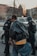 Three people are present, two of whom appear to be law enforcement officers wearing tactical gear and helmets. The third person, in the foreground, is wearing a leather jacket and carrying a bag. A vehicle is in the background with buildings and trees visible.