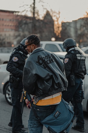 Three people are present, two of whom appear to be law enforcement officers wearing tactical gear and helmets. The third person, in the foreground, is wearing a leather jacket and carrying a bag. A vehicle is in the background with buildings and trees visible.