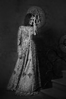 A person dressed in an intricately embroidered gown stands near a staircase. The gown features detailed floral patterns and is complemented by a matching choker. The background includes decorative circular designs, adding a touch of elegance to the scene.