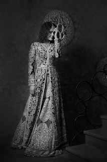 A person dressed in an intricately embroidered gown stands near a staircase. The gown features detailed floral patterns and is complemented by a matching choker. The background includes decorative circular designs, adding a touch of elegance to the scene.