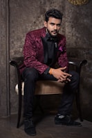 A man dressed in an elegant maroon jacket with floral patterns and black trim sits gracefully on a chair. He is wearing a black shirt, black bow tie, and black trousers, complemented by black shoes. The background features a textured, stone-like wall that adds a sophisticated ambiance.