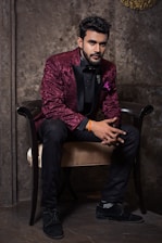 A man dressed in an elegant maroon jacket with floral patterns and black trim sits gracefully on a chair. He is wearing a black shirt, black bow tie, and black trousers, complemented by black shoes. The background features a textured, stone-like wall that adds a sophisticated ambiance.