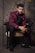 A man dressed in an elegant maroon jacket with floral patterns and black trim sits gracefully on a chair. He is wearing a black shirt, black bow tie, and black trousers, complemented by black shoes. The background features a textured, stone-like wall that adds a sophisticated ambiance.