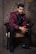 A man dressed in an elegant maroon jacket with floral patterns and black trim sits gracefully on a chair. He is wearing a black shirt, black bow tie, and black trousers, complemented by black shoes. The background features a textured, stone-like wall that adds a sophisticated ambiance.