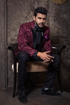 A man dressed in an elegant maroon jacket with floral patterns and black trim sits gracefully on a chair. He is wearing a black shirt, black bow tie, and black trousers, complemented by black shoes. The background features a textured, stone-like wall that adds a sophisticated ambiance.