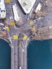 An aerial view of a busy urban intersection featuring multiple roads and crosswalks. Trams and cars are visible, along with buildings and a water body surrounding the area. The geometry of the roads and the arrangement of vehicles create an organized pattern.