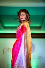 A person with curled brown hair and bright makeup is posing on a fashion runway. They are wearing a colorful saree with shades of red, pink, and gold. The background is softly lit with green lighting, adding a dramatic contrast to the attire.