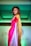 A person with curled brown hair and bright makeup is posing on a fashion runway. They are wearing a colorful saree with shades of red, pink, and gold. The background is softly lit with green lighting, adding a dramatic contrast to the attire.