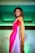 A person with curled brown hair and bright makeup is posing on a fashion runway. They are wearing a colorful saree with shades of red, pink, and gold. The background is softly lit with green lighting, adding a dramatic contrast to the attire.