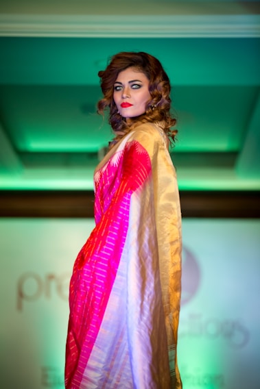 A person with curled brown hair and bright makeup is posing on a fashion runway. They are wearing a colorful saree with shades of red, pink, and gold. The background is softly lit with green lighting, adding a dramatic contrast to the attire.