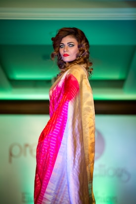 A person with curled brown hair and bright makeup is posing on a fashion runway. They are wearing a colorful saree with shades of red, pink, and gold. The background is softly lit with green lighting, adding a dramatic contrast to the attire.