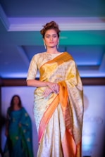 A woman is seen wearing an elegant gold and orange saree, adorned with intricate patterns. She has short, styled hair and is wearing dangling earrings. The background is illuminated with blue and green hues, indicating a fashion show runway setting with another individual visible in the distance.