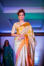 A woman is seen wearing an elegant gold and orange saree, adorned with intricate patterns. She has short, styled hair and is wearing dangling earrings. The background is illuminated with blue and green hues, indicating a fashion show runway setting with another individual visible in the distance.
