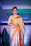 A woman is seen wearing an elegant gold and orange saree, adorned with intricate patterns. She has short, styled hair and is wearing dangling earrings. The background is illuminated with blue and green hues, indicating a fashion show runway setting with another individual visible in the distance.