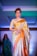 A woman is seen wearing an elegant gold and orange saree, adorned with intricate patterns. She has short, styled hair and is wearing dangling earrings. The background is illuminated with blue and green hues, indicating a fashion show runway setting with another individual visible in the distance.