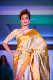 A graceful model wearing a flowing Benarasi saree with delicate floral embroidery, standing against a warm, traditional backdrop.