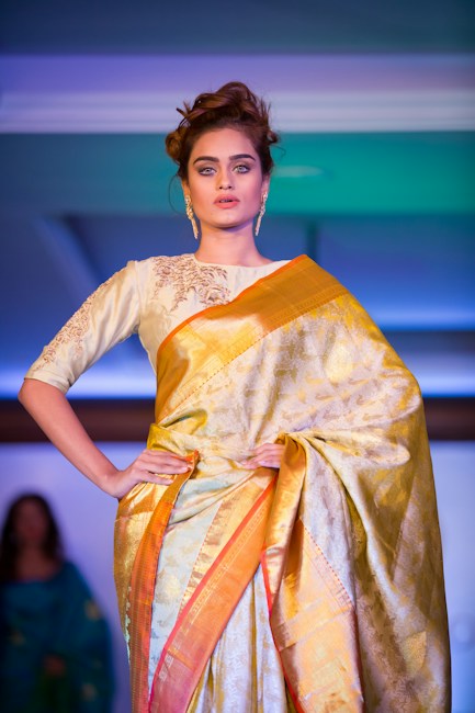 A woman stands confidently in a traditional attire, featuring a richly patterned silk saree with gold and orange hues. Her hair is styled in an elegant updo, and she wears ornate gold earrings. The background is softly lit with cool blue and green tones, creating a sophisticated and formal ambiance.