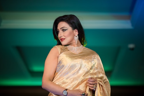 A woman wearing an elegant golden saree with intricate embroidery, large decorative earrings, and a watch on her wrist is caught in profile. The background features a soft gradient of green and blue tones, providing a rich contrast to her attire.