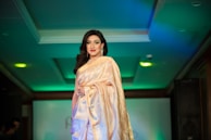 A person wearing a golden saree with intricate patterns, standing confidently. The setting appears to be a fashion event, surrounded by a dimly lit environment with green and blue lighting. The individual has dark hair and is adorned with earrings.
