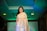 A person wearing a golden saree with intricate patterns, standing confidently. The setting appears to be a fashion event, surrounded by a dimly lit environment with green and blue lighting. The individual has dark hair and is adorned with earrings.