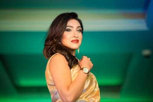 A woman is elegantly posing, wearing a gold dress and accessorized with a watch and earrings. Her hair is styled in soft waves, and she stands against a vibrant green background.
