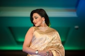 A woman in an elegant golden saree adorned with detailed embroidery, complemented by a matching blouse. She wears sparkling jewelry including a choker necklace and large earrings, and has a confident expression. Her dark hair is styled straight and she has accented eye makeup and red lipstick.