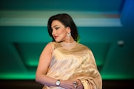 A woman in an elegant golden saree adorned with detailed embroidery, complemented by a matching blouse. She wears sparkling jewelry including a choker necklace and large earrings, and has a confident expression. Her dark hair is styled straight and she has accented eye makeup and red lipstick.