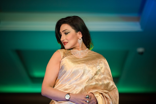 A woman in an elegant golden saree adorned with detailed embroidery, complemented by a matching blouse. She wears sparkling jewelry including a choker necklace and large earrings, and has a confident expression. Her dark hair is styled straight and she has accented eye makeup and red lipstick.