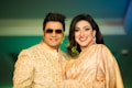 A smiling couple dressed in elegant traditional attire. The woman is wearing a gold sari with intricate detailing and the man is in a matching gold sherwani with a patterned design. Both exhibit a cheerful demeanor, with the woman sporting bold makeup and statement earrings. The background is softly blurred, casting a warm ambiance over the scene.