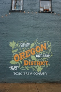 A mural painted on a brick wall features the text 'Welcome to the Oregon District' with an emphasis on 'Oregon' and 'District' in bold colors. The design includes decorative leaf motifs and mentions 'Dayton, Ohio' and 'Toxic Brew Company'. The wall has two upper windows and faintly seen string lights.