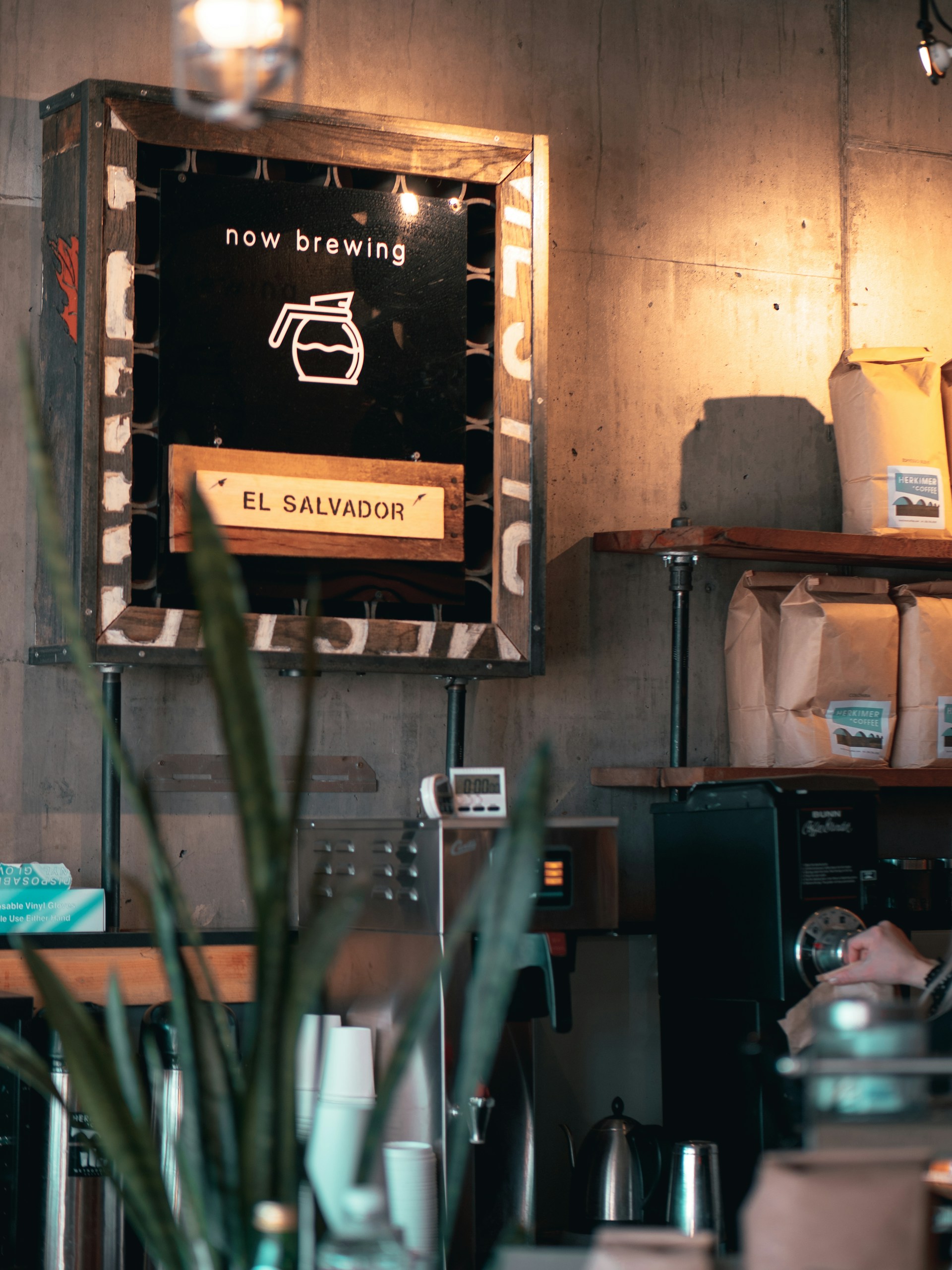 A cozy coffee shop interior featuring a rustic sign that reads 'now brewing' with a coffee pot icon and the words 'El Salvador' below. The background includes shelves with coffee bags, an espresso machine, and cups ready for use. The ambiance is warm, with soft lighting and a few plants adding a touch of greenery.