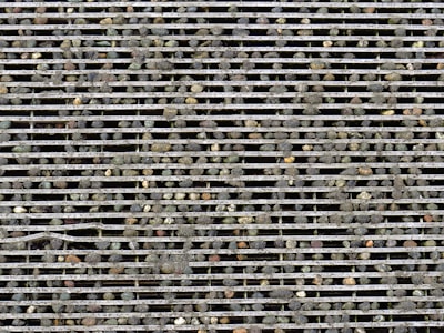 Rows of metallic bars form a grille with small pebbles scattered on and embedded between the bars, creating a rugged and textured surface.