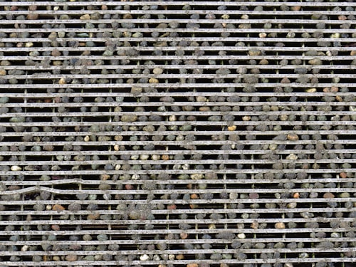 Rows of metallic bars form a grille with small pebbles scattered on and embedded between the bars, creating a rugged and textured surface.