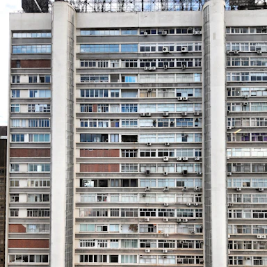 A multi-story residential building with a grid-like structure, featuring numerous windows and balconies. Many of the windows have air conditioning units installed, and the facade of the building is a mix of concrete, glass, and brickwork. The balconies are enclosed with glass or blinds, and some windows have curtains or drapes. The building's exterior shows signs of aging and varied maintenance levels.