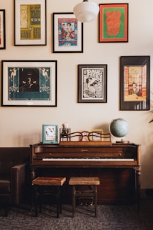 A room features a vintage upright piano with two wooden stools in front. On top of the piano, there are framed photographs and a globe. The wall above the piano is adorned with several framed posters of various sizes and colors. The decor suggests a cozy and artistic atmosphere.