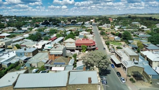 Aerial view of a small town with a main street lined with residential and commercial buildings. The surrounding area includes greenery and fields extending into the distance. The sky is partly cloudy, giving a bright and serene atmosphere.