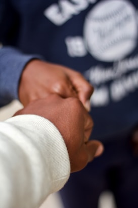 Two individuals are shown in close proximity, focusing on their hands clasped together, signifying connection or support. The background is blurred, highlighting a person wearing a dark sweatshirt.