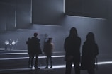 A dimly lit environment with several people standing and some walking. The setting is minimalistic and futuristic, with geometric lines of light illuminating the floor. The atmosphere is foggy, adding to the mysterious and abstract nature of the scene.