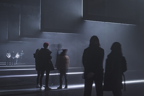 A dimly lit environment with several people standing and some walking. The setting is minimalistic and futuristic, with geometric lines of light illuminating the floor. The atmosphere is foggy, adding to the mysterious and abstract nature of the scene.