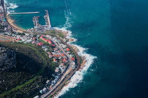 An aerial view of a coastal town with a prominent harbor protected by a breakwater. Houses with colorful rooftops are clustered near the shoreline, while lush green vegetation covers the area further inland. Waves from the ocean gently crash against the rocky shore, and a clear, expansive view of the deep blue sea stretches out on the right side.