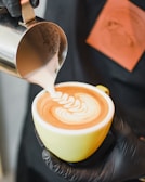 A barista is pouring steamed milk into a cup of coffee to create latte art, forming an intricate pattern on the surface. The cup is held in one hand, while the other hand pours the milk from a small metal pitcher.