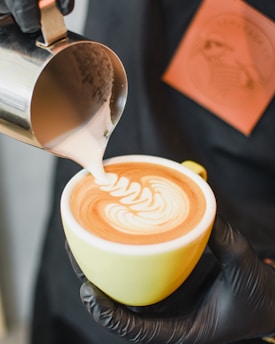 A barista is pouring steamed milk into a cup of coffee to create latte art, forming an intricate pattern on the surface. The cup is held in one hand, while the other hand pours the milk from a small metal pitcher.
