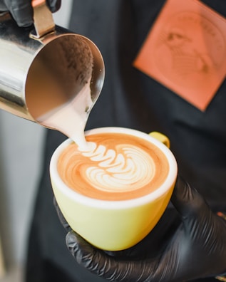 A barista is pouring steamed milk into a cup of coffee to create latte art, forming an intricate pattern on the surface. The cup is held in one hand, while the other hand pours the milk from a small metal pitcher.