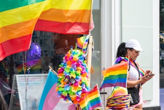 A person in a casual outfit stands beside a display adorned with various rainbow-themed items such as flags, banners, and a colorful flower lei. The person is looking at their phone while a large rainbow flag waves prominently in the background. A purple balloon can be seen in the reflection of the window.