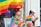 A person in a casual outfit stands beside a display adorned with various rainbow-themed items such as flags, banners, and a colorful flower lei. The person is looking at their phone while a large rainbow flag waves prominently in the background. A purple balloon can be seen in the reflection of the window.