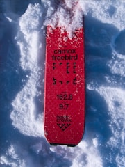 A red ski with the text 'camox freebird' covered partially with snow. The ski has specifications like 'length 162.8' and 'width 9.7' printed on it, along with the 'black crows' logo.