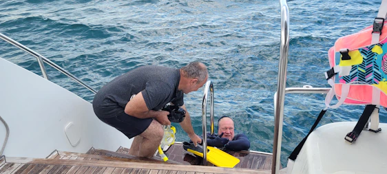Two men are interacting near the edge of a boat on water. One man is stepping down wooden stairs, holding underwater camera equipment. The other man, wearing a wetsuit, is in the water and holding a yellow flipper. A bright-colored life jacket is hanging on the right side rail. The ocean is calm with gentle waves visible in the background.
