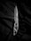 A silver folding knife is placed on a dark, textured fabric. The knife features an intricate cutout design on its handle and has a sharp, polished blade with a serrated edge near the base.