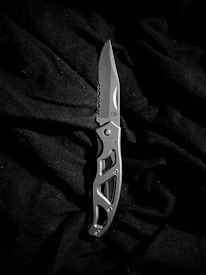 A silver folding knife is placed on a dark, textured fabric. The knife features an intricate cutout design on its handle and has a sharp, polished blade with a serrated edge near the base.